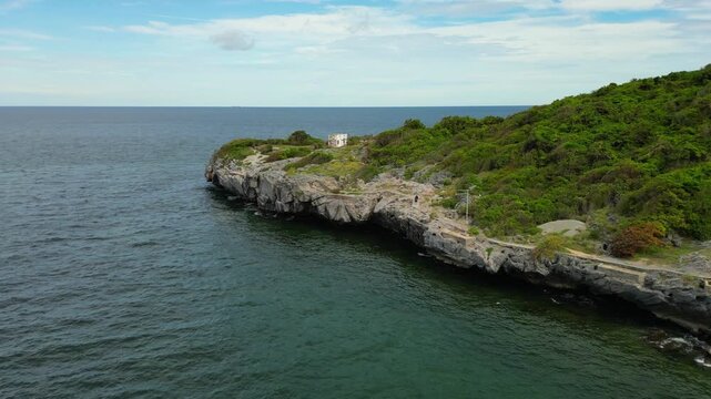 A cinematic drone shot captures a rugged, rocky coastline where a cliff covered in lush green trees meets the deep blue ocean. Gentle waves crash against the shore below. This stunning and peaceful fo