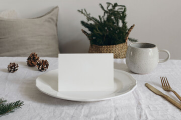 Festive Christmas table setting with blank place card on plate. Pinecones, gold cutlery, cup of coffee. Natural decor. Cozy rustic holiday. Greeting card mockup, seasonal design. Blurred background
