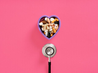 Stethoscope and heart shaped container full of pills lying on pink background