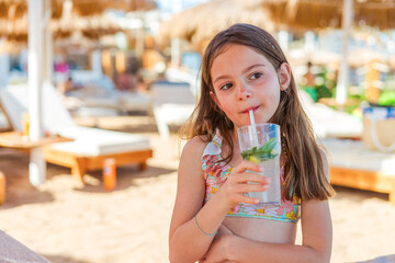 A cheerful child relaxes under beach umbrellas, sipping a cold nonalcoholic mojito in a tropical...