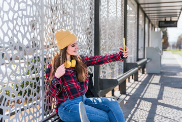 Happy female teenage in beanie taking a selfie with smartphone while waiting at transport stop