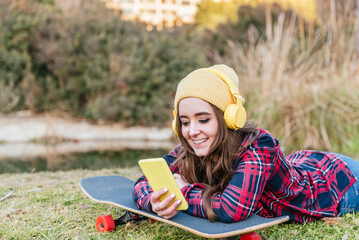 Teenage girl in yellow beanie lying on skateboard with headphones using smartphone in park