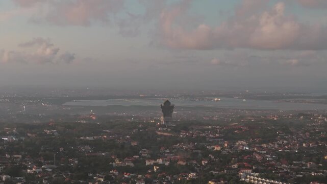 Aerial view glides toward Garuda Wisnu Kencana monument above Denpasar and the Bali Mandara toll bridge at golden dusk, with pink cloud and city light forming a calm horizon ideal for travel branding