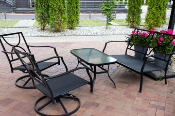 Outdoor seating area with black metal chairs, glass table and bench near flower pots on paved terrace