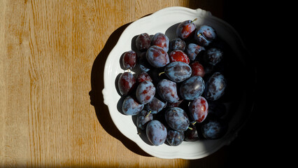 Close-up of fresh plums in a white ceramic bowl on a wooden surface. Summer harvest illuminated by soft natural light