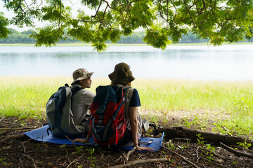 Man and woman hikers resting under tree near lake and meadow.