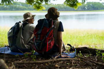 Man and woman hikers resting under tree near lake and meadow.