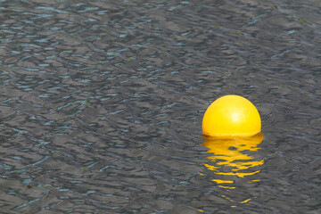 Bright yellow buoy floating on calm rippling water, creating reflection and contrast against dark...