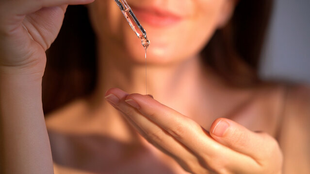 Young woman holding a dropper with serum for face. Facial oil fall from a glass pipette, macro view. Selective focus. Cosmetic background with liquid skincare product, beauty concept. Dermatology