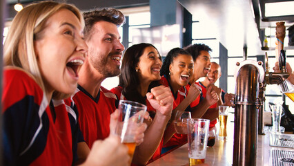 Multi-Cultural Group Of Friends Wearing Team Shirts In Sports Bar Celebrating Watching Game On TV
