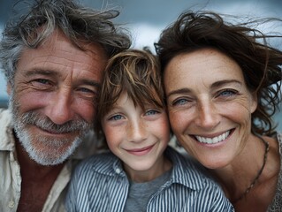 A smiling family of three poses closely together outdoors with the ocean visible in the background above them.