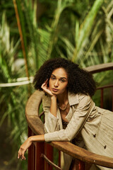 Stylish woman with curly hair poses in tropical setting in trench coat