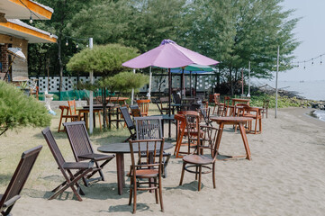 Relaxing beachside cafe setup with wooden chairs, tables, umbrellas, and ocean view under sunny sky, perfect summer holiday vibe