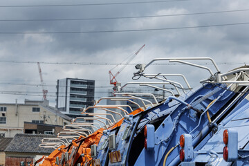 Immaculately clean garbage trucks line parked during weekend down time. White control arms sit open above truck's colorful light-blue or orange bodies. Scene blends industry and city life elements.