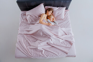 A mother and her daughter are nestled comfortably in bed, enjoying a quiet morning together under...