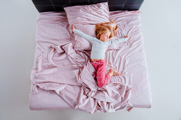 young girl is curled up in a ball on a bed with pink sheets and pillows. The bright, airy room has...