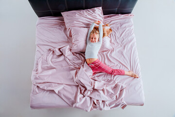 young girl is curled up in a ball on a bed with pink sheets and pillows. The bright, airy room has...