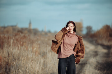 Teenage girl with red hair and glasses in a rural field, making a peace sign, dressed warmly in autumn.