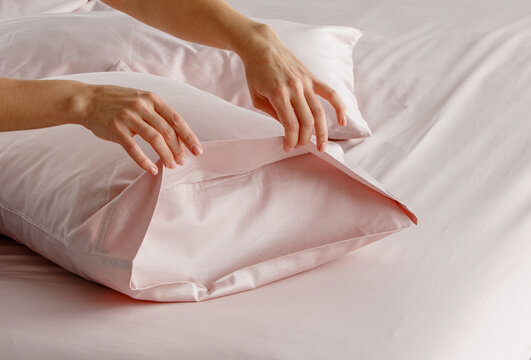 A hand gently pulls back a silky pillow cover in a soft pink bedding arrangement, creating an inviting atmosphere in a modern bedroom setting during daylight.