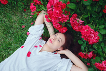 Teenage girl with red hair lying on grass, surrounded by red roses, hands behind head, wearing...