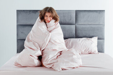 A woman with wavy hair sits on a neatly made bed, wrapped in a light pink blanket, radiating a calm...