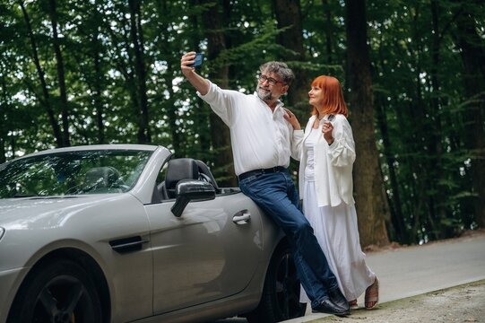 Doing the selfie. Lovely senior couple standing on the road near convertible cabriolet car - Powered by Adobe