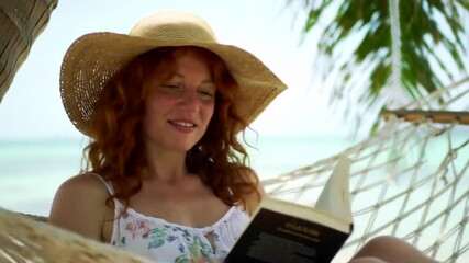 Relaxing on a tropical beach hammock, a woman enjoys reading a book under the sun by the serene coastline.