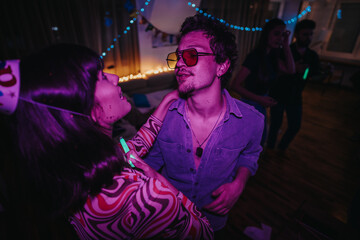 Two young adults share a joyful moment at a vibrant birthday party. The room is adorned with festive lights, creating an energetic atmosphere as other guests celebrate in the background.