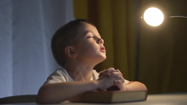 Young boy is praying. Religious leader prays to a lifestyle child. Book of prayers on a table in the evening. A boy is offering a prayer.