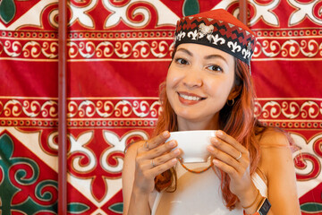 Portrait of smiling Kazakh woman wearing traditional headdress holding a cup of tea inside a yurt...