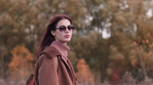 Teen girl smiling softly at the camera, dressed warmly in a brown coat and sunglasses, set against a fall landscape.
