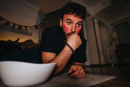 A young man lies on the floor with face paint, feeling hungover after a lively birthday party. A bowl is nearby, and a birthday banner is visible in the background.