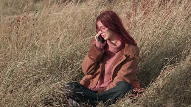 Teen girl with glasses calmly talking on her phone, sitting in tall grass, wearing a warm brown coat and pink sweater in a serene autumn field.