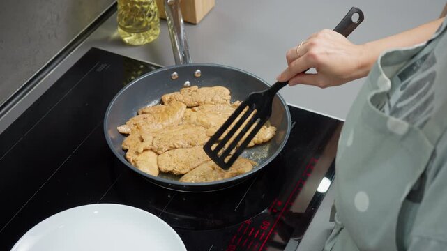 A woman is cooking marinated chicken fillets in a hot non-stick pan on an induction stove. She flips the meat with a spatula and covers the pan with a glass lid to cook it through.