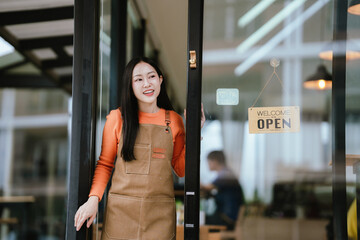 Smiling female barista in apron standing at café entrance, giving thumbs up, welcoming customers with joy and confidence.