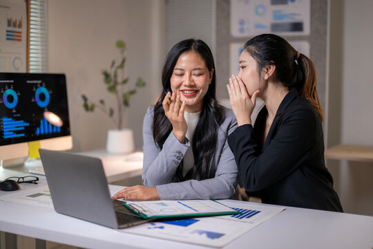 Businesswomen sharing secrets while analyzing charts on laptop in office