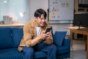 Obraz premium Asian freelancer enjoying using smartphone on sofa at home office
