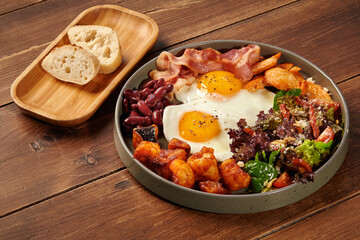Delicious breakfast consisting of fried eggs, bacon, beans, chicken, potatoes and salad on wooden table