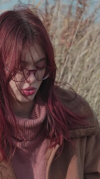Teen girl in a close-up with wind-blown hair across her face, wearing a pink sweater in a natural autumn scene.