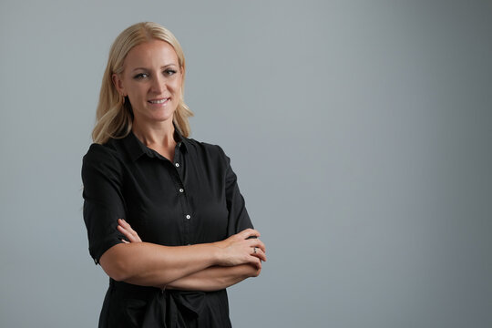 Portrait of a happy confident business woman in black shirt standing with arms crossed isolated on grey background, looking at the camera and smiling.