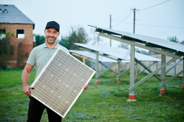 Man holds solar panel while standing in solar farm. Happy guy wearing black cap and light green...