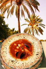 Fish dish soup top view. Close-up of freshly cooked European fish soup served on a plate with Mediterranean Sea palms background. Healthy food concept.