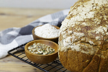 Detail of a loaf of bread on a rack next to two wooden bowls filled with seeds. The bread is toasted and has a crispy crust. Sourdough bread.