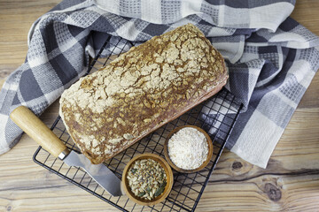 Flat lay of a loaf of whole wheat bread on a cooling rack next to two bowls, one with flour and one with sunflower seeds, and a knife. It's on a wooden table.
