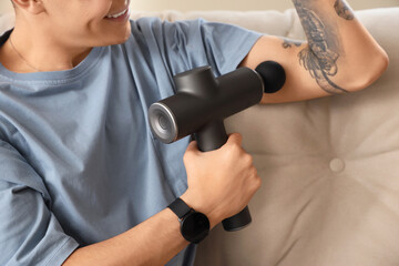 Fototapeta premium Young man massaging his arm with percussive massager at home, closeup
