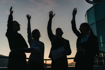 A group of business people silhouetted against a sunset backdrop, raising hands in a dynamic and...