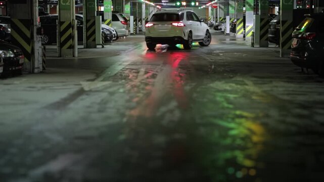 White Suv car Drives Away Through Dimly Lit Underground Garage, Taillights Reflecting On Wet Surface. Dark Hatchback Stops and Turns in background