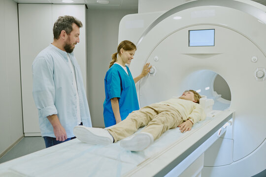 Caucasian boy lying on MRI scanner bed preparing for medical imaging procedure, young Caucasian woman technician operating machine, middle aged Caucasian man standing nearby observing