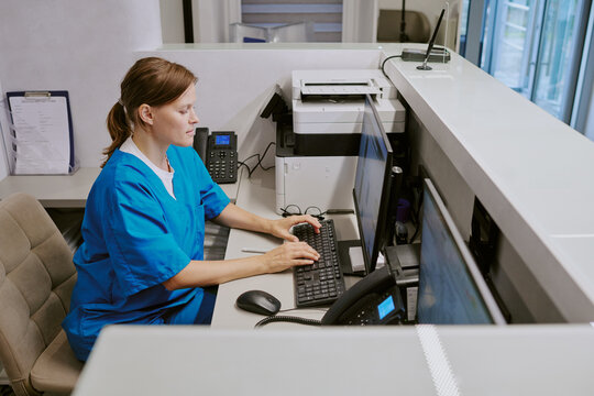 Caucasian middle aged woman working at medical reception desk typing on computer keyboard, sitting in modern healthcare facility, using office equipment and telephone