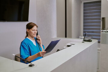 Caucasian young adult woman sitting at reception desk talking on phone while holding clipboard,...